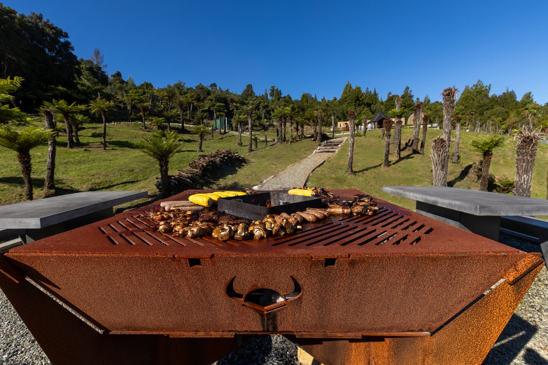 Corten steel barbecue grilling corn, sausages and mushrooms with barrel cabins and tree ferns beyond
